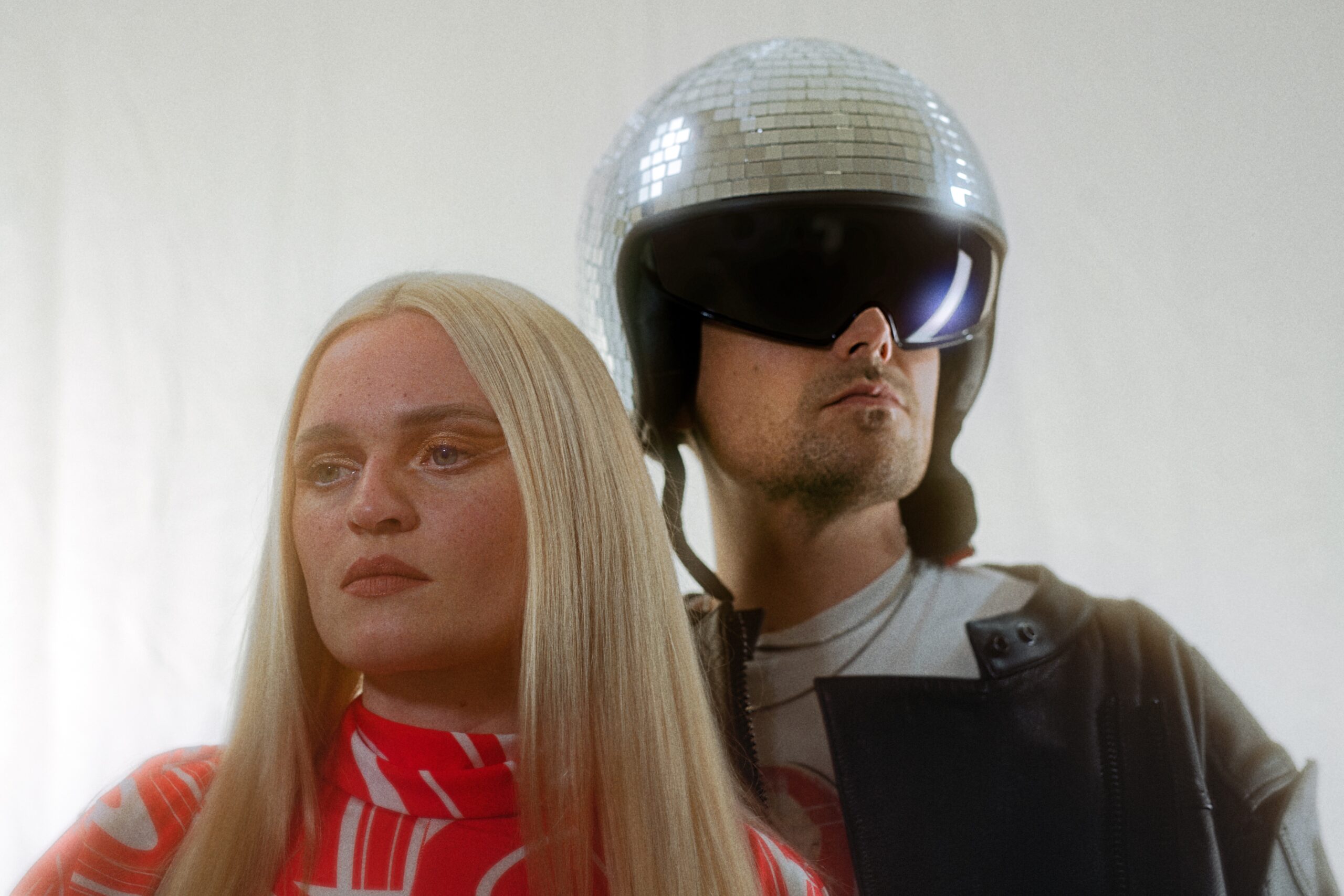 Die beiden stehen eng nebeneinander, Inéz blickt ernst, Demian trägt eine riesige Sonnenbrille und einen Helm, der wie eine Diskokugel aussieht.