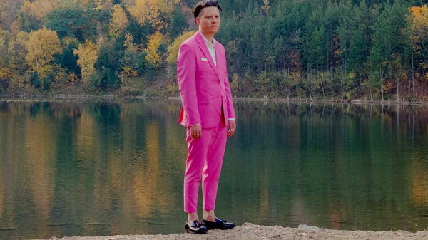 Konstantin Gropper steht am Ufer eines Sees, im Hintergrund ist Wald zu sehen. Er trägt einen sehr pinken Anzug. Mit Einstecktuch