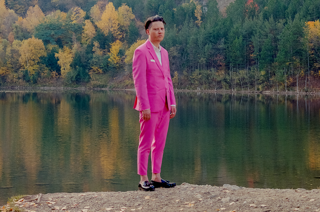 Konstantin Gropper steht am Ufer eines Sees, im Hintergrund ist Wald zu sehen. Er trägt einen sehr pinken Anzug. Mit Einstecktuch