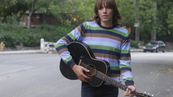 Evan Dando trägt einen gestreiften Pulli, läuft eine Straße entlang und hält eine schwarze Akustikgitarre in den Händen