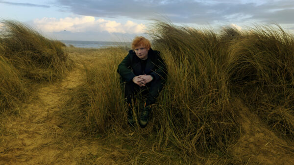 Sheeran sitzt auf einer Düne, Wind weht, bewölkter Himmel, hinter ihm das Meer