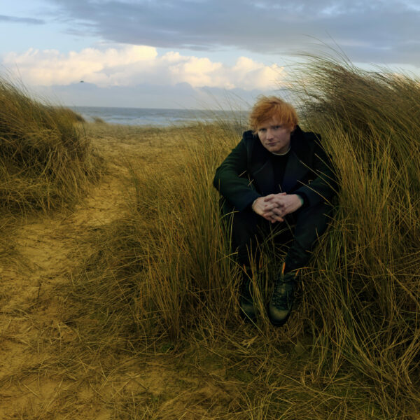 Sheeran sitzt auf einer Düne, Wind weht, bewölkter Himmel, hinter ihm das Meer