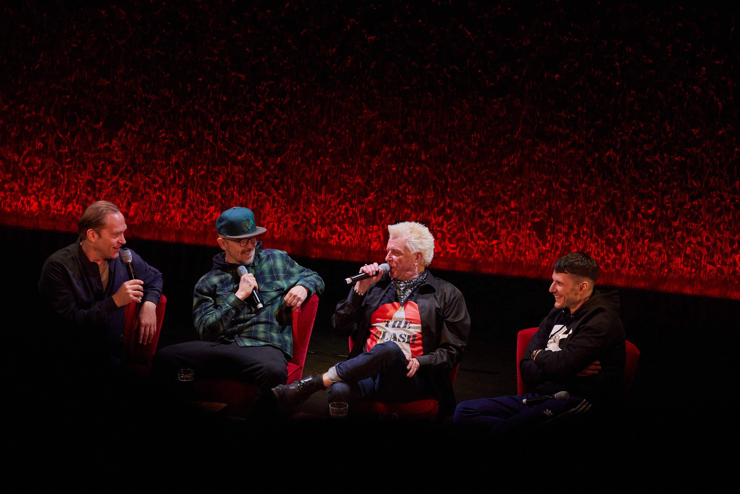 Vier Männer auf der Bühne, sie sitzen auf Sesseln vor einem roten Vorhang, sprechen und lachen