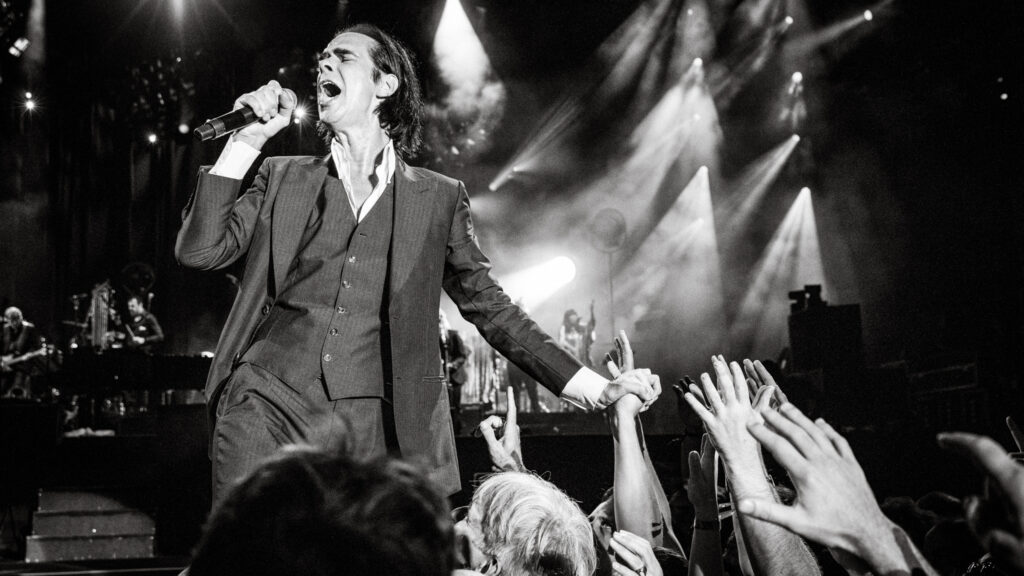 Schwarzweißfoto, Nick Cave singt ins Mikrofon, mit der anderen Hand hält er die Hand eines Fans