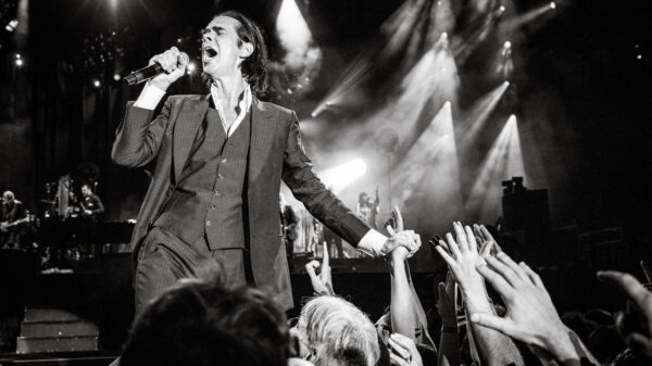 Schwarzweißfoto, Nick Cave singt ins Mikrofon, mit der anderen Hand hält er die Hand eines Fans