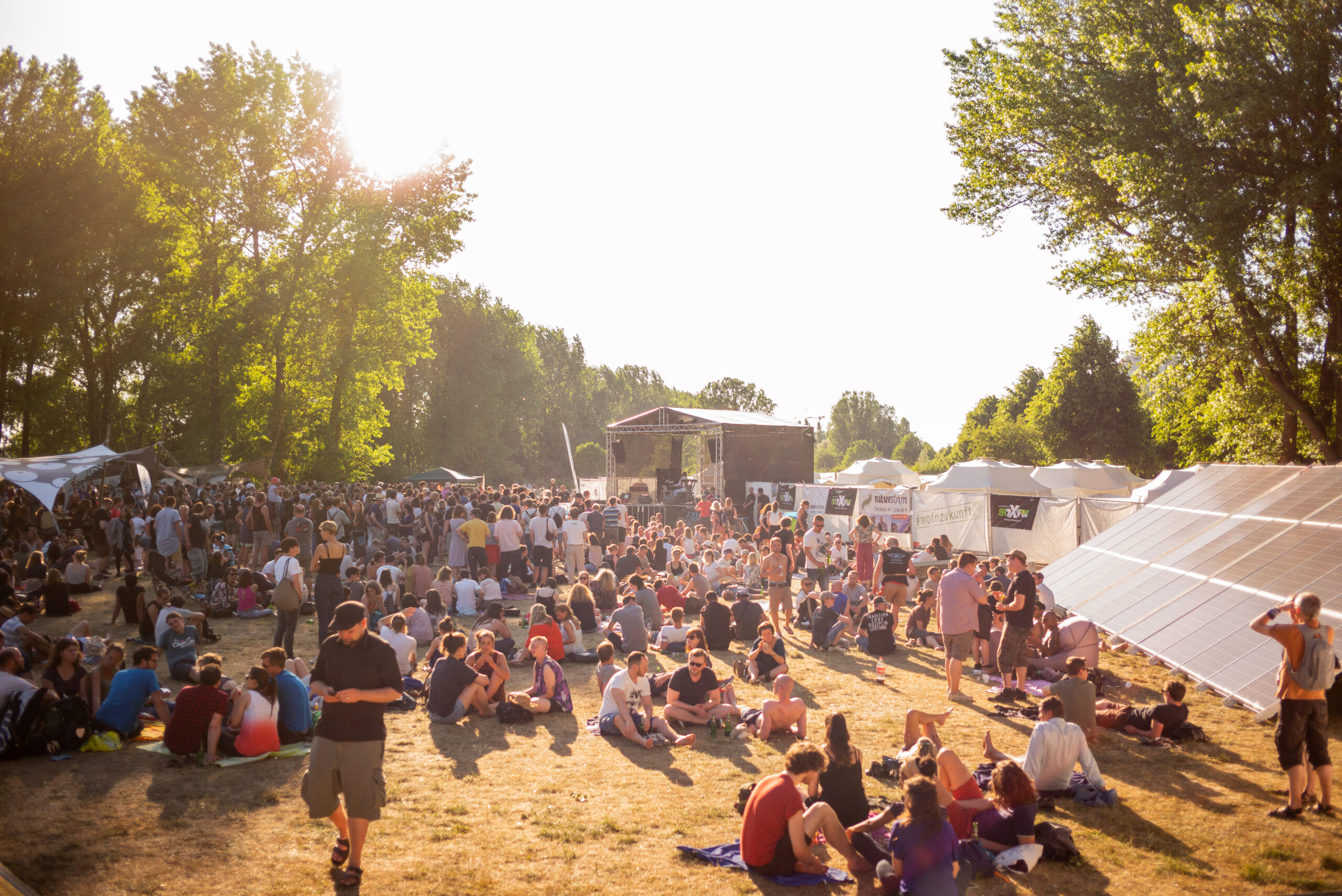 Elbpark bei Sonnenschein, rechts Solarpeanele, hnten eine Bühne, davor sitzen und stehen Menschen