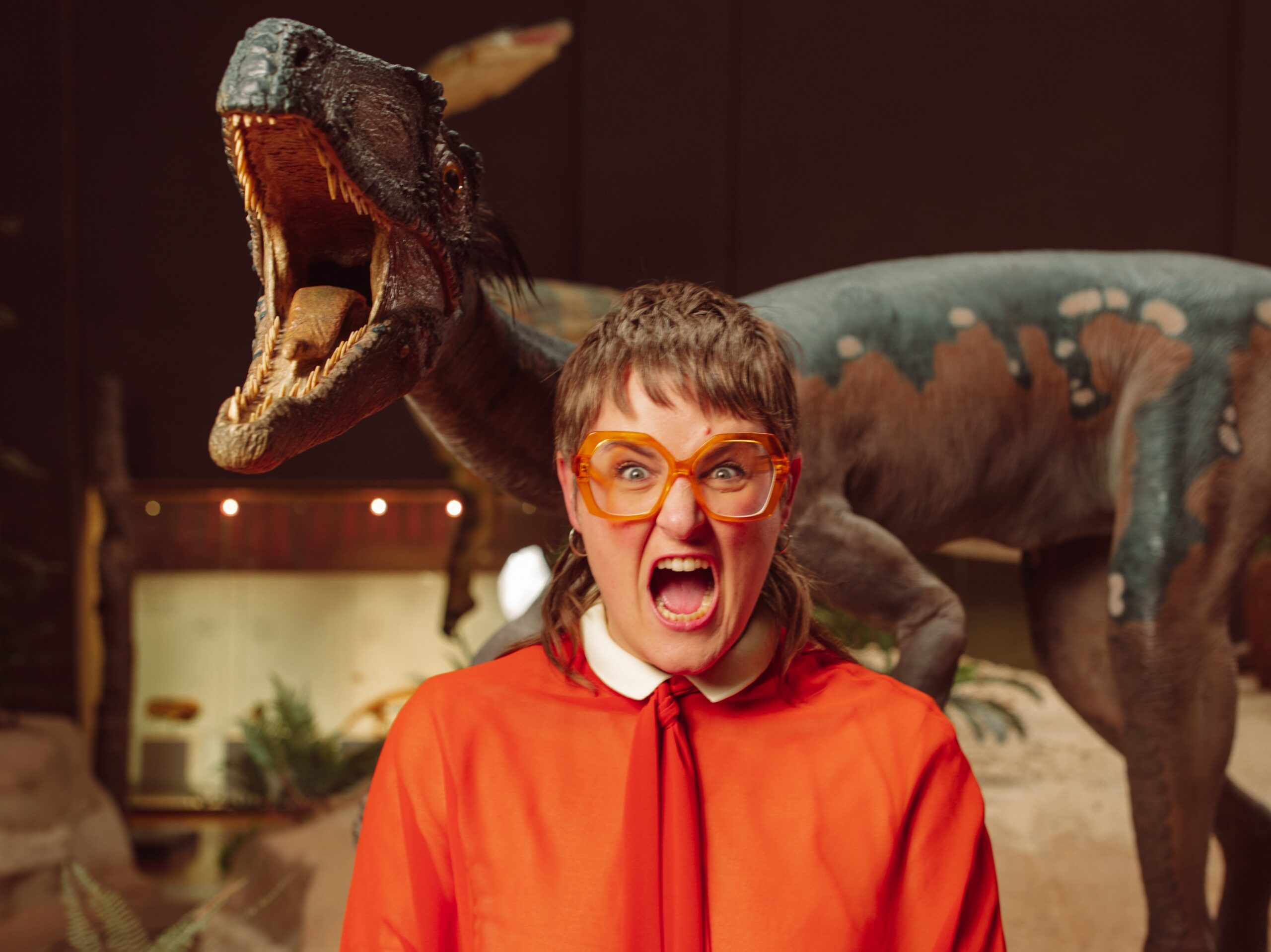 Eine junge Frau, kurze braune Haare, rote Brille, weite rote Bluse zieht eine Grimasse, hinter ihr eine Dinosaurier-Figur