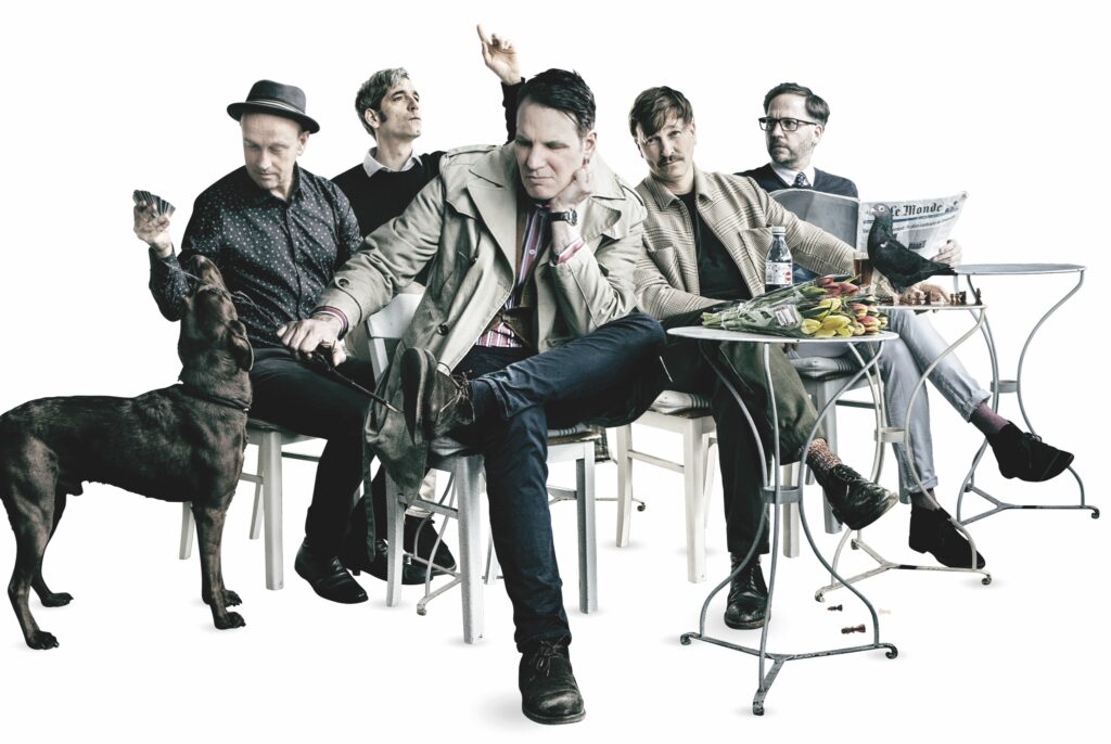 Bandfoto, die fünf Männer sitzen an Cafétischen, ein Hund ist auch im Bild