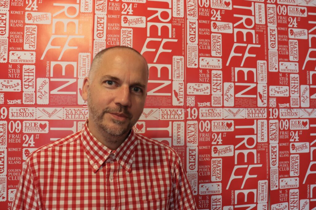 Er steht vor einer einem rot-weißen Hintergrund, der aus den Plakaten des Festivals besteht