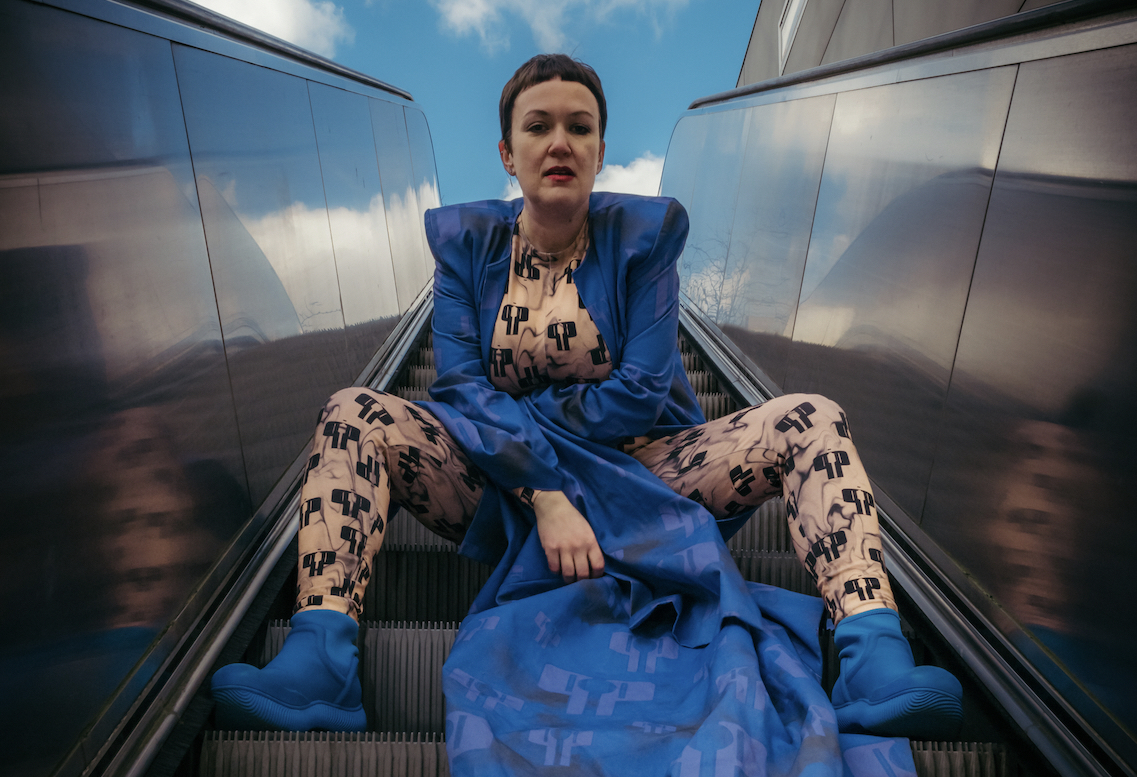 Sie sitzt in einem blauen Kleid und mit blauen Schuhen auf einer Rolltreppe