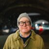 Er steht draußen, im Hintergrund vielleicht ein Tunnel, er trägt Jacke, Brille und Mütze