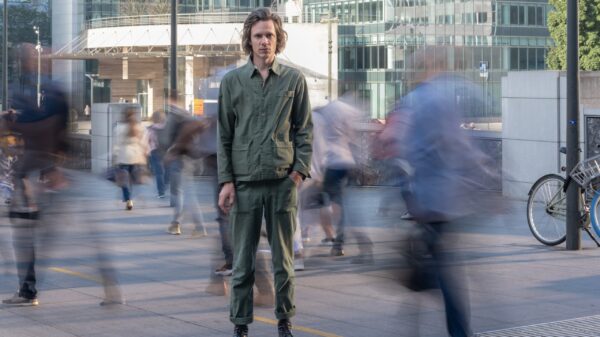 Er steht draußen auf einem Platz, die Menschen, die sich um ihn herum bewegen, sind bewegungsunscharf.