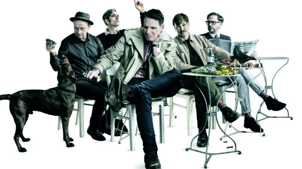 Die Band sitzt an Café-Tischen, einer liest Zeitung, links ist ein Hund zu sehen.