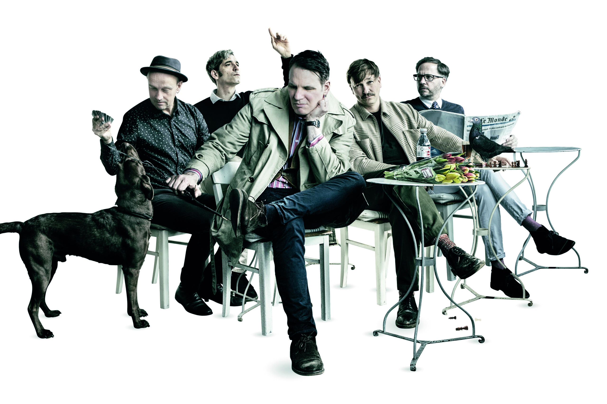 Die Band sitzt an Café-Tischen, einer liest Zeitung, links ist ein Hund zu sehen.