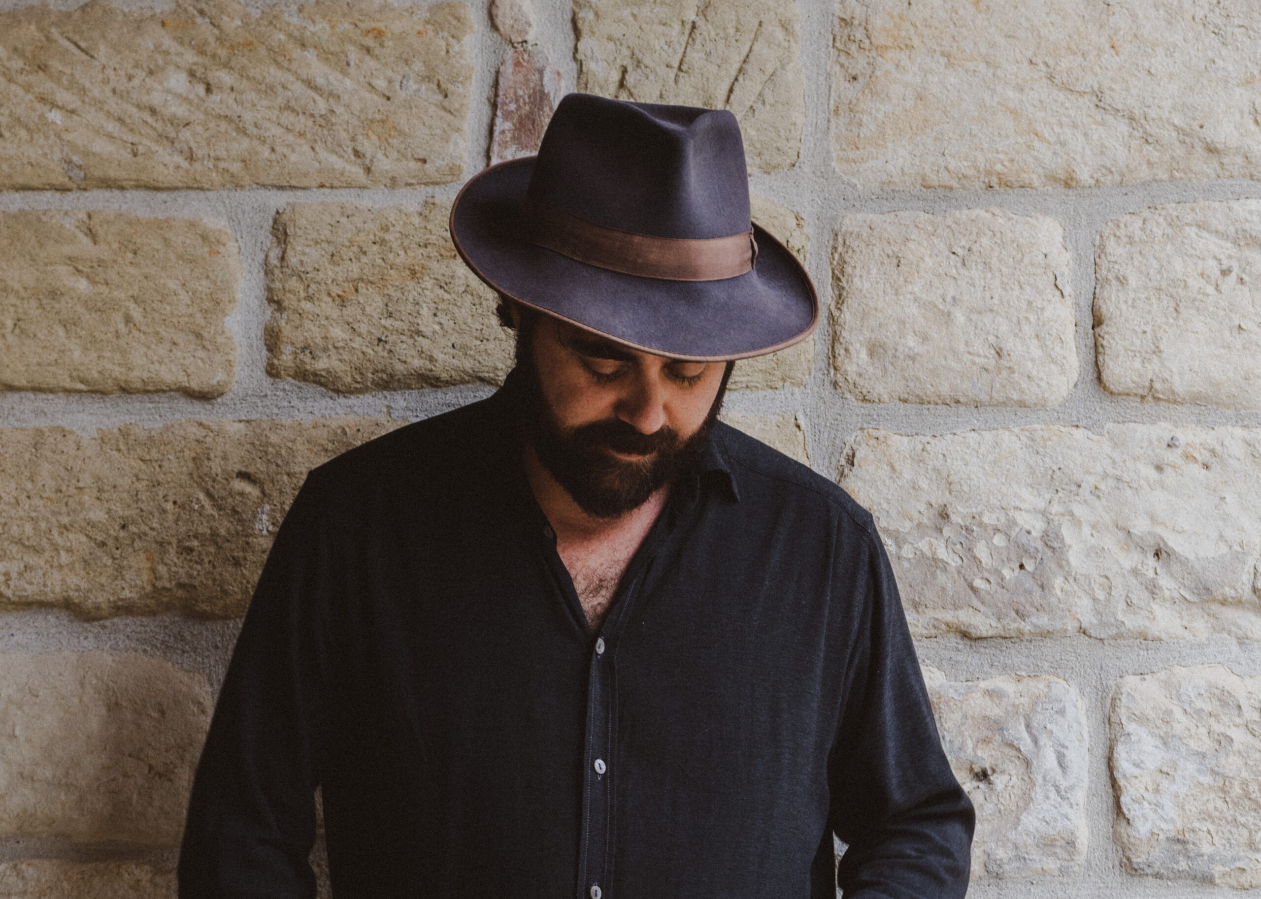 Der Musiker steht vor einer Wand, schaut nach unten, er trägt ein dunkles Hemd und einen Hut
