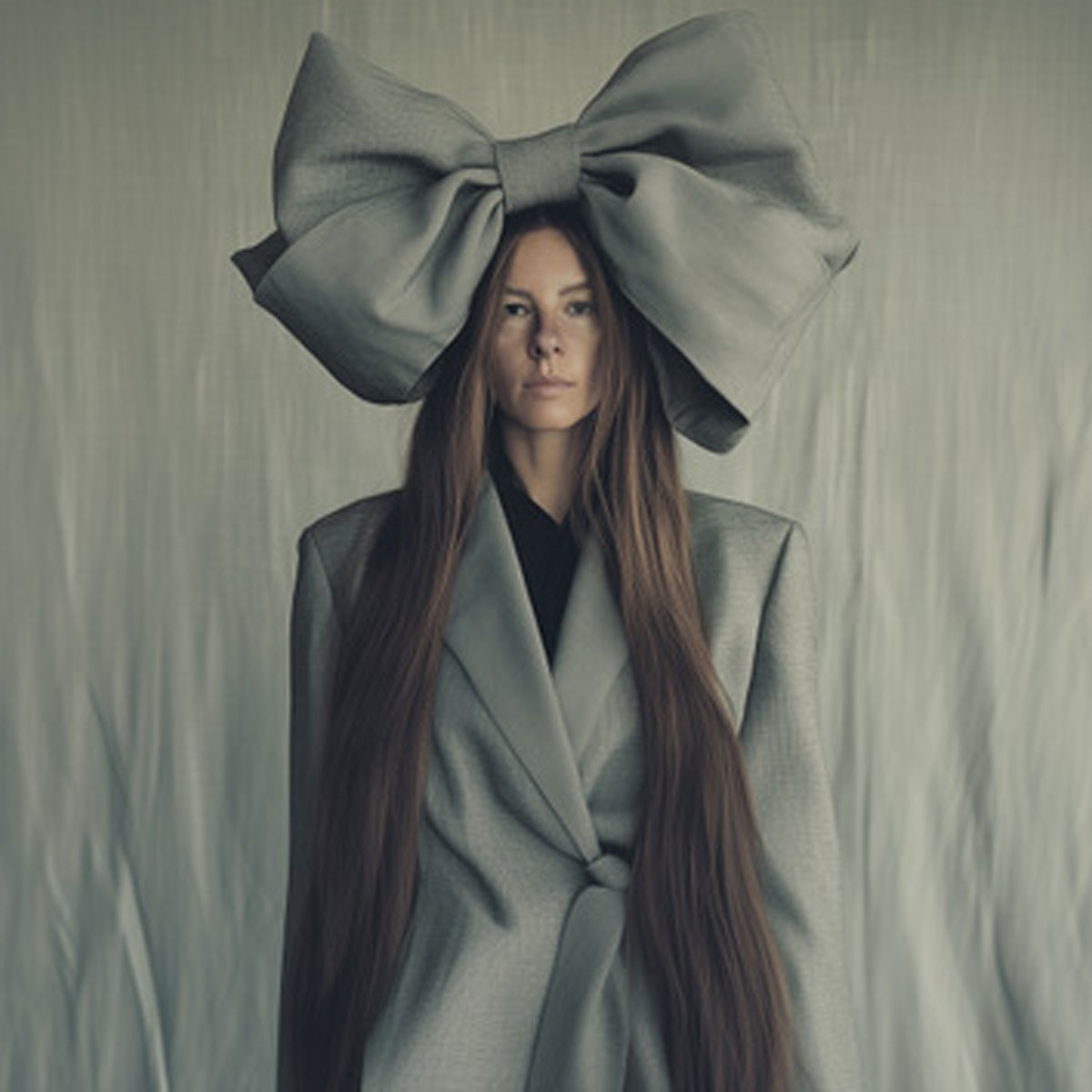 Die Künstlerin im grauen Kostüm vor einer grauen Wand, auf dem Kopf eine riesige graue Schleife