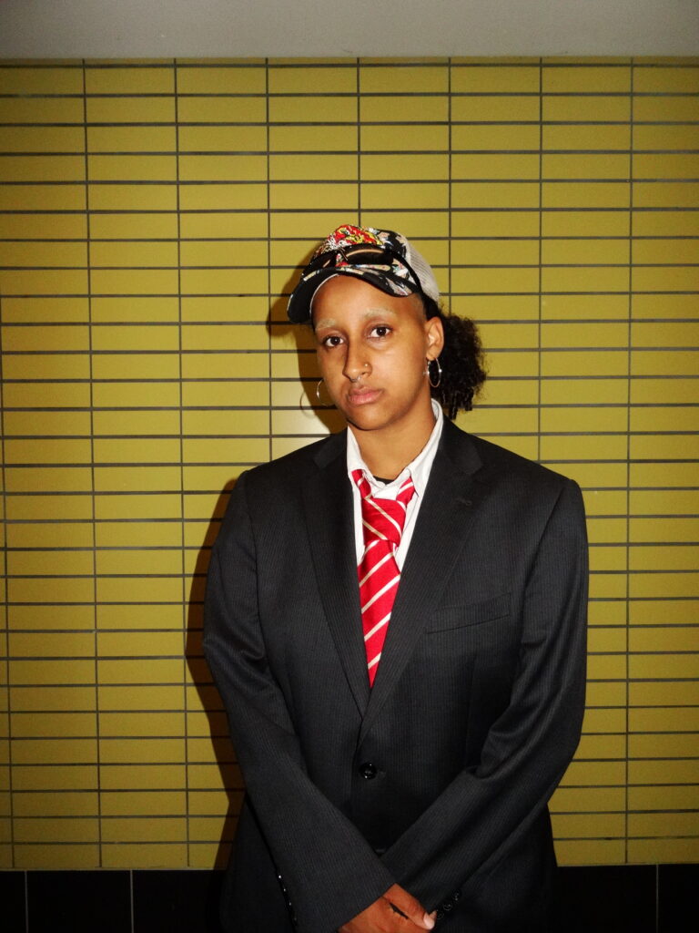 Steht vor einer gelben Kachelwand und schaut in die Kamera