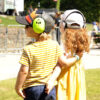 Zwei kleine Kinder stehen bei Sonnenschein Arm in Arm vor der Stadtpark-Bühne, beide mit Gehörschutz und Caps. Im Hintergrund weitere Menschen und die Bühne