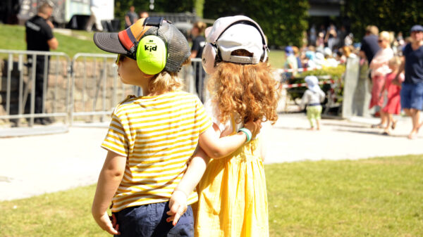 Zwei kleine Kinder stehen bei Sonnenschein Arm in Arm vor der Stadtpark-Bühne, beide mit Gehörschutz und Caps. Im Hintergrund weitere Menschen und die Bühne