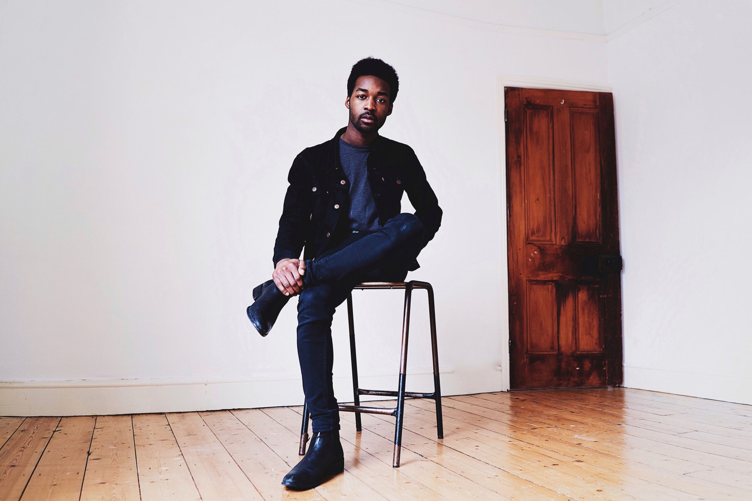 Er sitzt auf einem Hocker in einem sonst eher leeren Raum