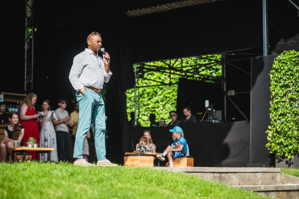 Er steht auf der Bühne, im Hintergrund sieht man Menschen stehen und sitzen