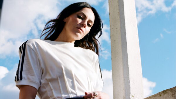 Die Sängerin trägt eine Art Fußball-Trikot und lehnt sich an ein Geländer, hinter ihr blauer Himmel mit Wolken