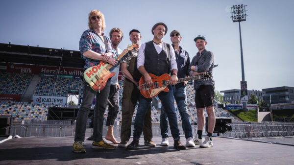 Sechs Männer stehen - teils mit Gitarren - auf einer Bühne in einem leeren Stadion