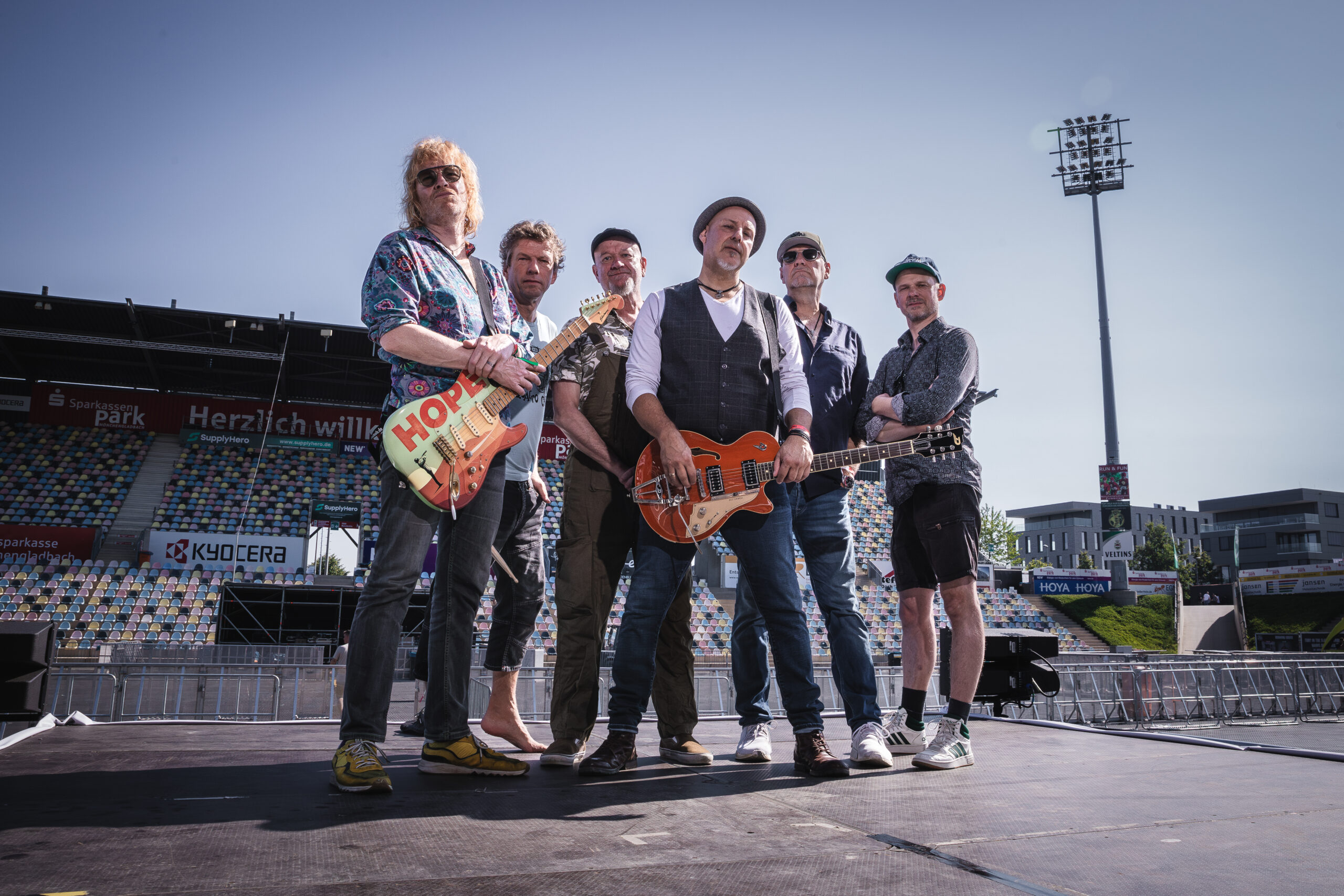 Sechs Männer stehen - teils mit Gitarren - auf einer Bühne in einem leeren Stadion
