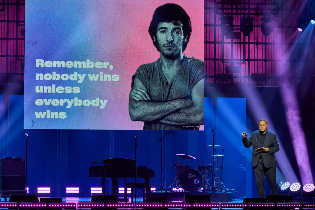 Brosda auf der Bühne, im Hintergrund auf der Leinwand Springsteen und die Worte „Remember, nobody wins unless everybody wins“