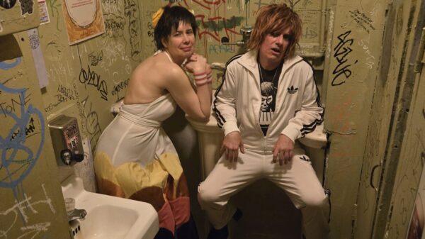 Ein Mann und eine Frau, beide tragen weiße Kleidung, sitzen auf einer Toilette, Aufkleber und Schriftzüge an den Wänden