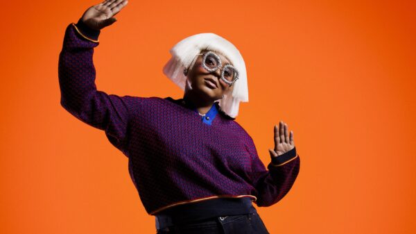 Sie steht vor einem orangefarbenen Hintergrund, gestikuliert, trägt eine weiße Perücke und eine auffällige Brille