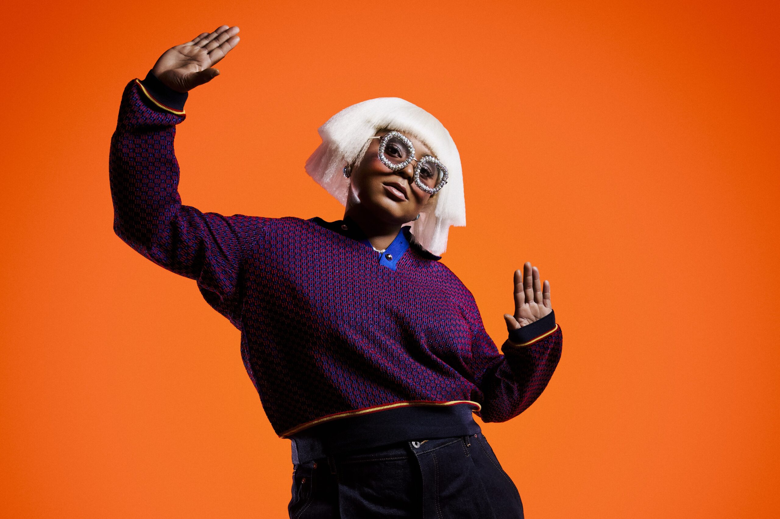 Sie steht vor einem orangefarbenen Hintergrund, gestikuliert, trägt eine weiße Perücke und eine auffällige Brille