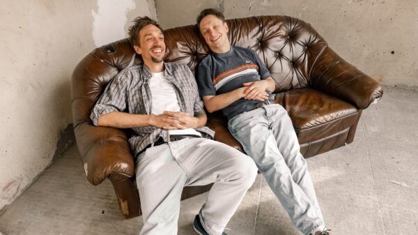 Zwei Männer hängen auf einem braunen Ledersofa ab, das in einer Ecke steht. Beide lächeln.