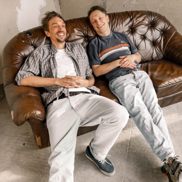 Zwei Männer hängen auf einem braunen Ledersofa ab, das in einer Ecke steht. Beide lächeln.