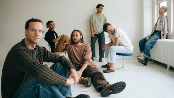 Die Band – sechs Männer - in einem weißen Raum, sie sitzen auf dem Boden, auf Stühlen, auf einem Heizkörper oder lehnen an der Wand