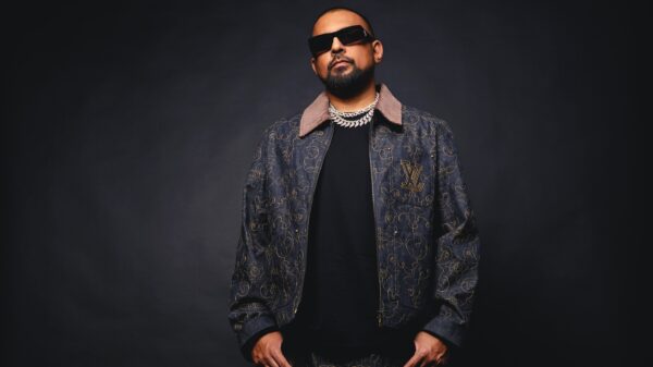 Er steht vor einer dunklen Wand, trägt Sonnenbrille, hat die Daumen in den Hosentaschen und blickt ernst in die Kamera