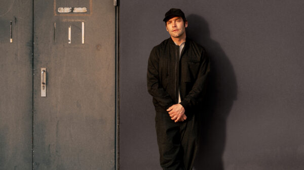 Bosse steht - in schwarzer Kleidung und mit Cap - vor einer dunklen Wand und schaut in die Kamera.