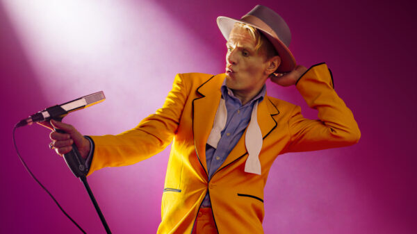 Alexander Scheer steht in einem orangefarbenen Anzug an einem Mikrofon.