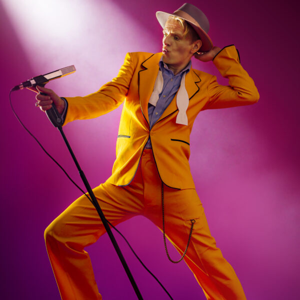 Alexander Scheer steht in einem orangefarbenen Anzug an einem Mikrofon.