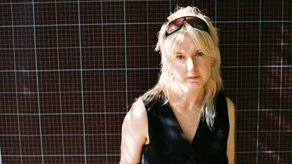 Sie steht vor einer Wand, Sonnenbrille im Haar und schaut in die Kamera.