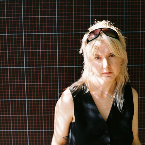 Sie steht vor einer Wand, Sonnenbrille im Haar und schaut in die Kamera.