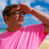 Mann mit Brille und einem pinken T-Shirt steht am Strand.
