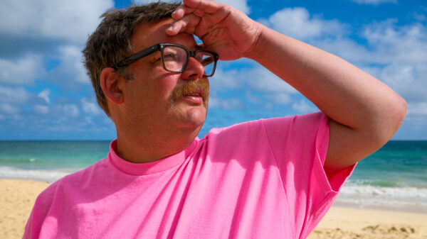 Mann mit Brille und einem pinken T-Shirt steht am Strand.