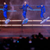 Die Backstreet Boys tanzen in blauen Outfits auf einer beleuchteten Bühne vor jubelndem Publikum.