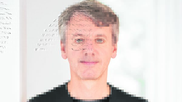 Frontalporträt eines Mannes mit schwarzem Oberteil vor hellem Hintergrund.