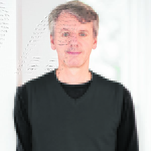 Frontalporträt eines Mannes mit schwarzem Oberteil vor hellem Hintergrund.
