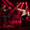Musiker spielen mit Gitarren und Schlagzeug auf einer rot beleuchteten Konzertbühne.