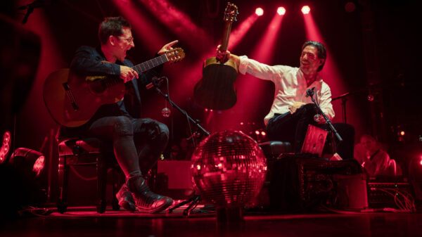 Musiker spielen mit Gitarren und Schlagzeug auf einer rot beleuchteten Konzertbühne.