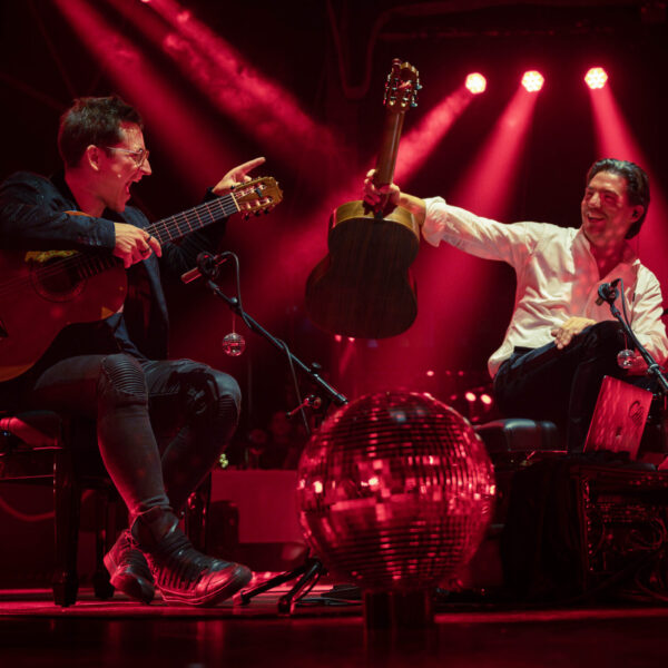 Musiker spielen mit Gitarren und Schlagzeug auf einer rot beleuchteten Konzertbühne.